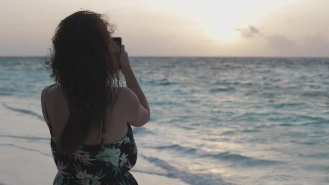 Back view of woman holding smartphone and Take Photo on the sunset at the Maldives beach