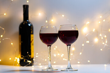 Two transparent glasses filled with red wine stand next to each other. A bottle of red wine, two filled glasses, a bokeh of lights in the background.