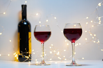 Two transparent glasses filled with red wine stand at a distance from each other. A bottle of red wine, two filled glasses, a bokeh of lights in the background.