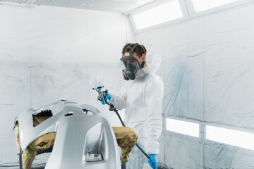 Car painter in a protective suit and mask varnishes a painted bumper of a vehicle while working in...