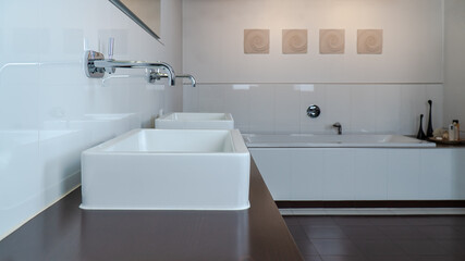 Fototapeta na wymiar Modern bathroom in white and brown with two sinks, bathtub and wooden furniture. Selective focus on the front