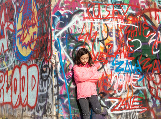 Moody pensive emotional young girl teenager with long dark hair in a pink jacket standing in front of a graffiti wall.