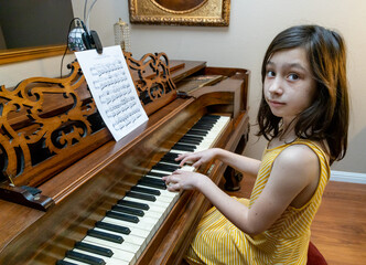 A cute clever young girl with delicate fingers and coy mona lisa smile during piano practice playing a rare antique Chickering Square Grand piano built-in 1867 wearing a yellow dress. - obrazy, fototapety, plakaty