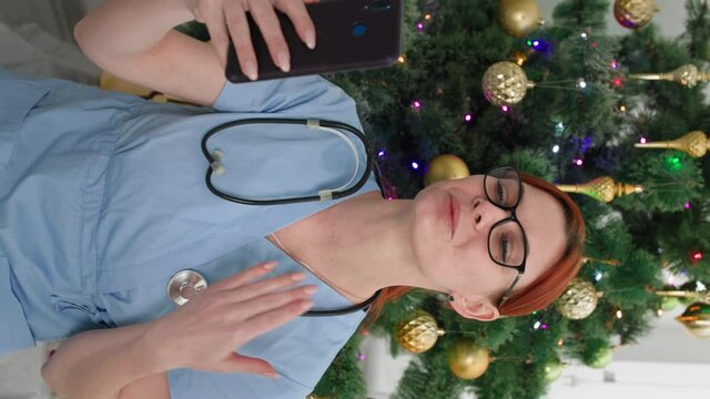 celebration via video link, young female doctor communicates with family on video camera on smartphone and congratulates on Christmas holidays sitting near the Christmas tree, verti