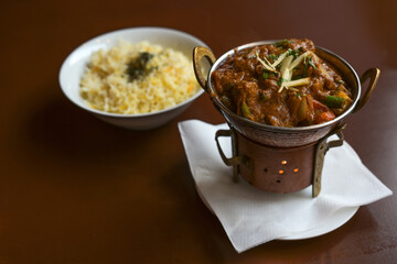 Indian curry dish with meat, vegetables and ginger served on a small metal stove with a candle like a tea warmer and a bowl of rice on a brown table, copy space, selected focus