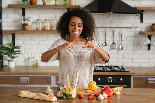 Vlogging blogging concept. African young woman teenage girl cooking vegetable vegan salad while taking photo for social media on smart phone online in the kitchen.