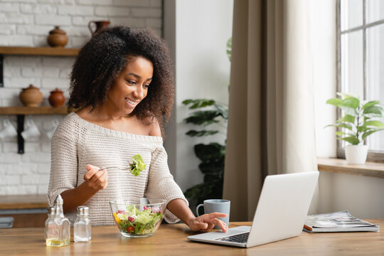 Smiling african young woman girl eating vegan vegetarian salad at home kitchen while watching webinars online on laptop, working on distance, e-learning from home