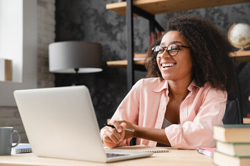 Cheerful laughing young african woman tutor freelancer student writing at copybook homework test,...