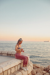 Fototapeta na wymiar Full-length photo of asian lady in sportswear sitting on the seashore in the evening, opening a bottle of water