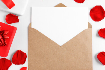 Open envelope with list on white wooden table with red rose petals and gift boxes top view. Mockup for greeting card, invitation, flat lay.