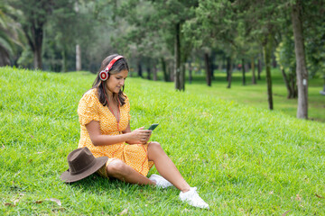Woman sitting on the grass listening to music with headphones while texting with her cell phone