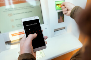 Person using mobile phone with verification code at an ATM. Security concept for authentication by...