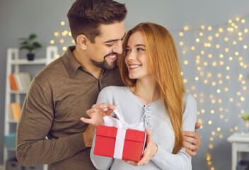 Fototapeta Christmas, birthday, anniversary, holiday present concept. Young smiling woman opening gift box with ribbon from loving hugging husband or boyfriend at home with decorated home interior at background obraz