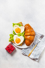 Two heart-shaped eggs with lettuce and croissant on a grey background. Romantic breakfast with red gift box. Beautiful Valentine's Day meal service