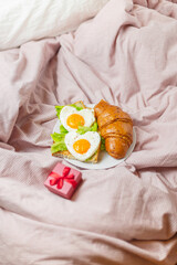 Fototapeta na wymiar Heart-shaped scrambled eggs with salad and croissant on the bed. Romantic breakfast in bed. Beautiful meal delivery by February 14th. Breakfast and red gift