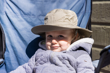 Closeup front view of a cute two year old caucasian boy, sitting outside on a deckchair in the backyard wearing a cotton hat with copy space to left.
