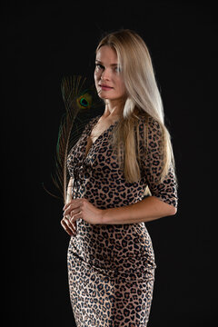 Young girl stands on black background and in her hands she holds peacock feather. She is blonde with long hair.