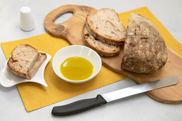 Fresh bread with crispy crust on wooden cutting board with with olive oil in the bowl and salt, knife and orange napkin. Fresh Sliced sourdough bread loaf, tasty appetiser.
