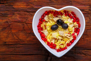 Puff salad with chicken breast and pineapple, garnished with pomegranate and olives in a heart salad bowl on a wooden table.