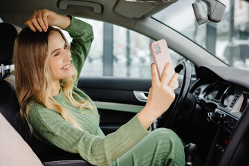 Horizontal shot of good looking female poses for selfie in smart phone, sits at wheel in luxurious car,  and shares photos in social networks, uses free internet connection
