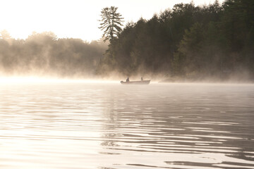Obraz na płótnie Canvas Early morning fisherman in a boat on a misty lake with evergreen trees in the background