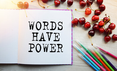 The inscription WORDS HAVE POWER in a notebook. View of the table along with scattered cherries and colored pencils. Flat layout. Conceptual photography.
