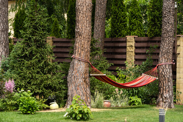 Red hammock swing in metal frame with nobody on green lawn in backyard near log house cottage. Rest relax relaxation alone on hammock swing in Summer garden. Long web banner.