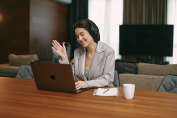 Smiling young woman in headphones waving hand, greeting, welcoming chatting, make video call, looking at laptop screen