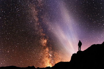 Silhouette of a person on the top of mountain in starry night sky.  Bright milky way galaxy behind him.