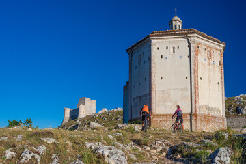 Man and woman riding mtb to castle ruins on mountain top at Rocca Calascio, italian travel destination, Gran Sasso National Park, Abruzzo, Italy. Clear blue sky