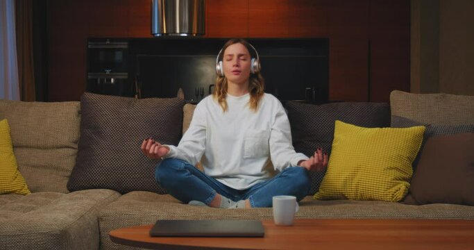 Happy calm young woman in headphones listen to relaxing music for yoga meditation, sitting in lotus pose on sofa at home