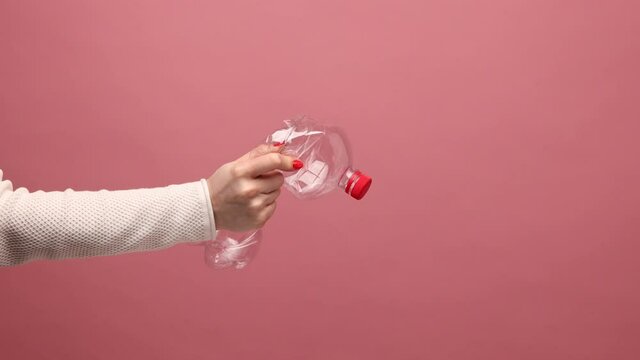 Profile portrait of woman hand holding and pressing plastic bottle with red cap, sorting his rubbish, worrying about environmental pollution. Indoor studio shot isolated on pink background.