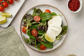 Salad with green leaves, lettuce, spinach, cherry tomatoes, mozzarella, olive oil, chilli pepper, lime juice on grey stone surface, top view