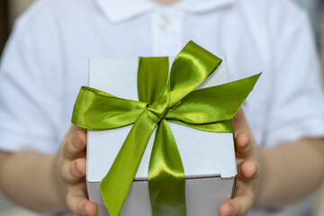 holding a white gift box with green color ribbon in his hands. cute kid in white t shirt is smiling or has serious face. free space, for greeting card, womans or mother's
