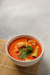 White bowl of fresh Tom Yum soup on a bamboo stand on a light background