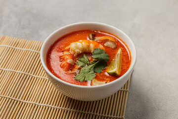 White bowl of fresh Tom Yum soup on a bamboo stand on a light background