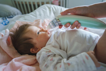 Side view on unknown woman mother using electric baby nasal aspirator mucus nose suction sucking the saliva from baby's nose cleaning while lying on the bed baby with atopic eczema - obrazy, fototapety, plakaty