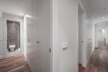 Fototapeta na wymiar Long hallway with cabinet lined walls, corner cloakroom with open door and white built-ins with dark wood floor
