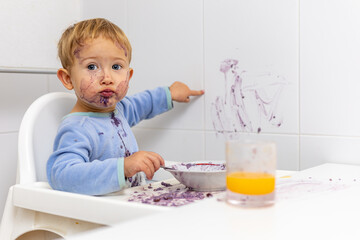 Niño rubio con la cara manchada señala la pared que acaba de manchar con su comida