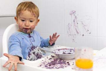 Un niño rubio de 2 años con pijama azul sentado en una trona con la cara sucia muestra la mano...