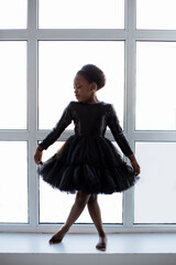 African american little kid wearing stylish black dress standing in some ballet pose near window at studio. Tender little ballerina with bare feet posing indoors. © sofiko14