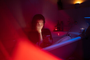 Young woman with mental disorder, alcohol addiction or relationship problems sit in clothes in bath with cold water reading messages from ex-boyfriend. Exhausted girl suffer from depression illness