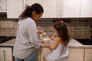 Rear view of charming girl mixing dry ingredients in a glass bowl and her mother pouring some vanilla powder into flour while making pancake dough in home kitchen. Mother and daughter cooking together