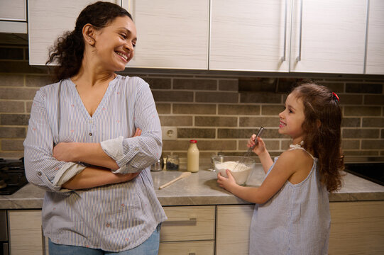 Beautiful smiling woman, happy loving mother leaning at kitchen countertop and admiring her adorable daughter cute baby girl mixing ingredients in a glass bowl while learning kneading pancakes dough