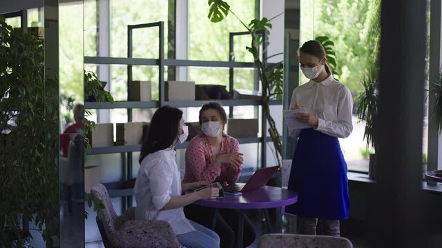 Wide shot women sitting at table in cafe ordering in slow motion as waitress in coronavirus face mask standing writing order with pen. Positive Caucasian clients indoors in coffee house on pandemic