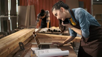 Young Asian female carpenter using laptop computer while talking to customer on the phone