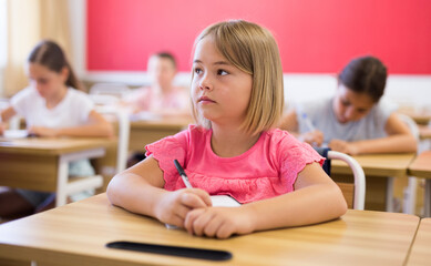 Fototapeta na wymiar Schoolgirl sitting at desk in classroom during lesson. Her classmates sitting around.