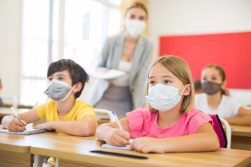 Fototapeta premium Portrait of cute little schoolgirl in protective face mask studying in classroom. Necessary precautions in coronavirus pandemic..