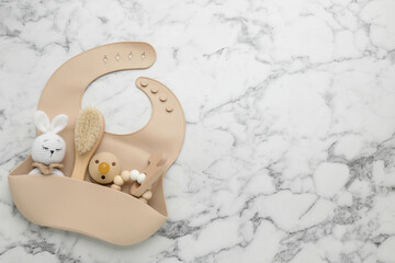Flat lay composition with silicone baby bib, toys and accessories on white marble background. Space...