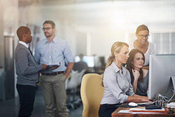 Business as usual. Shot of three women working on a computer with colleagues blurred in the background.